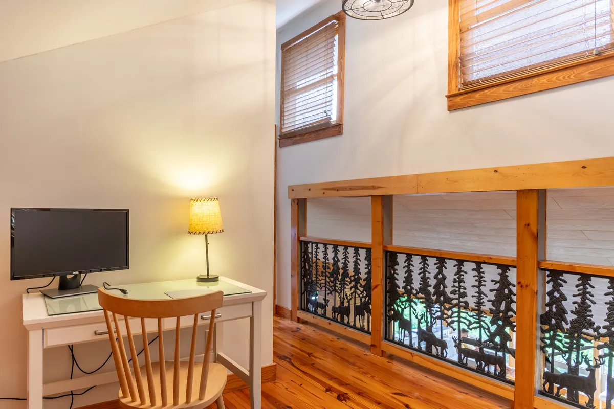 A tranquil loft area with a desk, computer, table lamp, and a decorative wooden railing with cutouts. The space is well-lit and organized, ideal for remote work, creative pursuits, or journaling in peace.