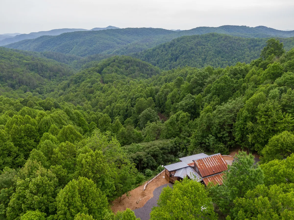 A breathtaking view image highlighting a wide mountain range, lush green hills, and the cabin with its metal roof nestled among the trees. The scene evokes a tranquil, expansive sense of place and connects directly to the experience of exploring and being immersed in mountain beauty.
