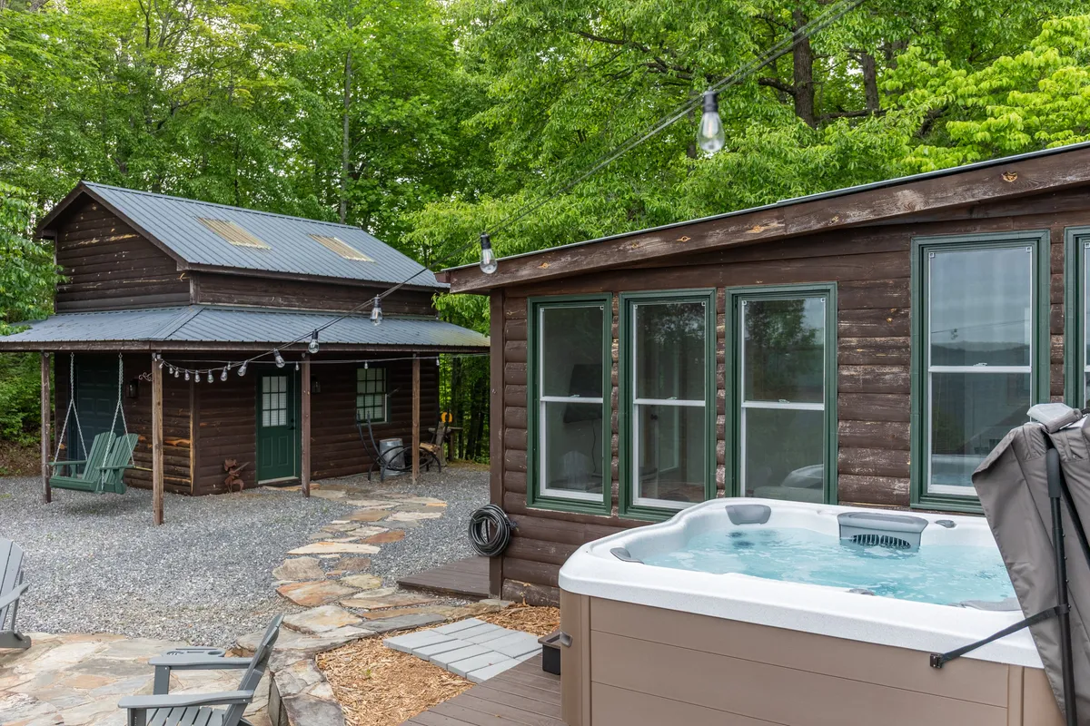 Here, the exterior of the wooden cabin is featured with a hot tub, a swinging chair, and a landscaped stone pathway. The scene exudes relaxation and connection with nature, providing a glimpse of outdoor amenities perfect for breaks between work sessions.