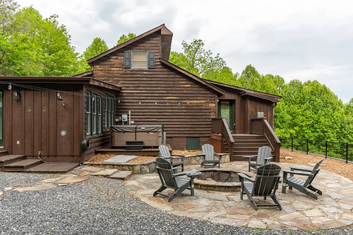 A welcoming exterior shot showcasing the wooden cabin with a stone patio, fire pit, hot tub, and abundant surrounding greenery. The scene highlights communal outdoor amenities and the inviting atmosphere for gatherings.