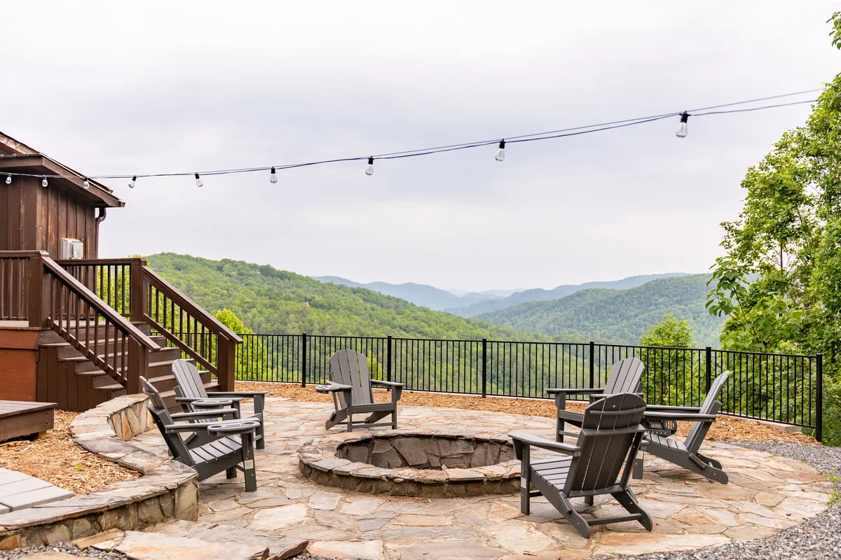 A welcoming outdoor setting featuring a stone fire pit surrounded by Adirondack chairs, string lights overhead, and a breathtaking mountain view in the background. The area is ideal for gathering, with a cozy and inviting ambiance for both day and night.