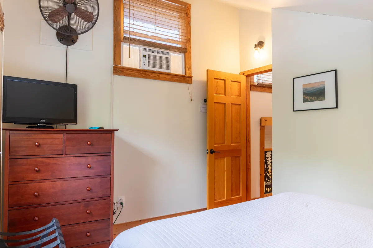 A serene bedroom featuring a king-sized bed, a wooden dresser, a TV, and a doorway, bathed in natural light. The room's calming decor and comfortable furnishings invite restful nights and peaceful mornings.