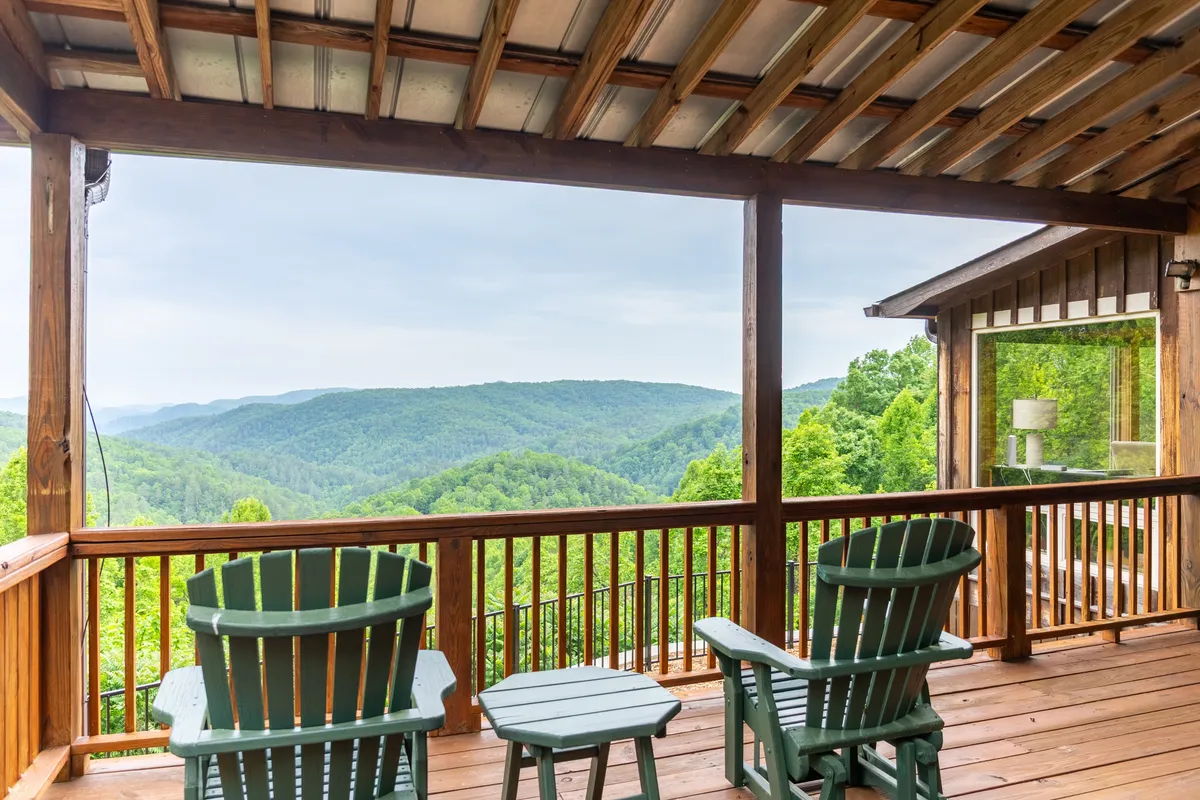 A spacious wooden deck with Adirondack chairs arranged to face panoramic mountain views. The glass wall of the house enhances the connection between indoors and outdoors, suggesting a space ideal for reading, meditating, or family cookouts.