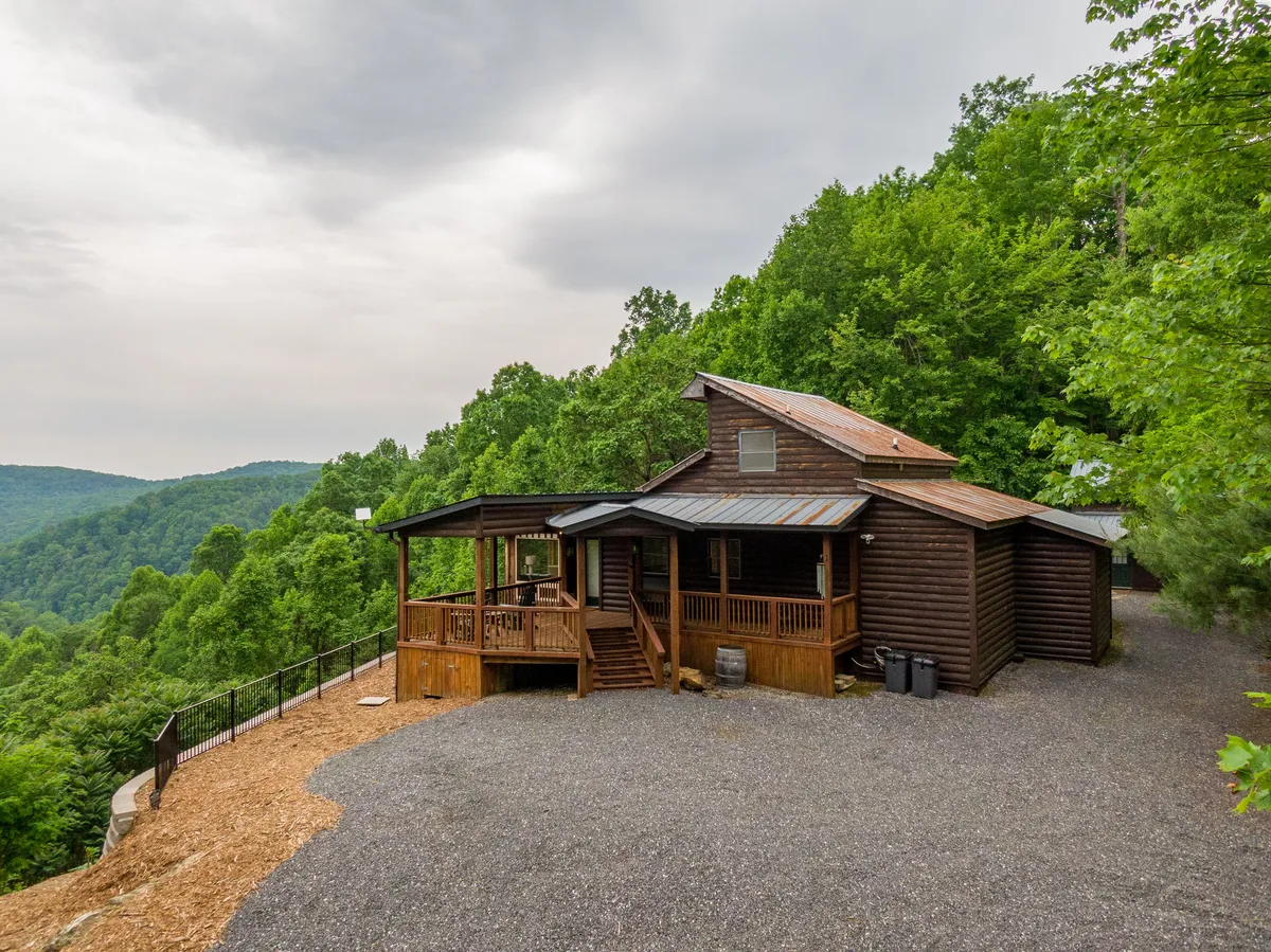 A serene scene showing a log cabin surrounded by lush greenery, with a spacious deck area and a gravel driveway leading up to the property. The deck provides a perfect spot for morning coffee or evening stargazing, and the wide open space emphasizes the cabin's seclusion in the mountains.