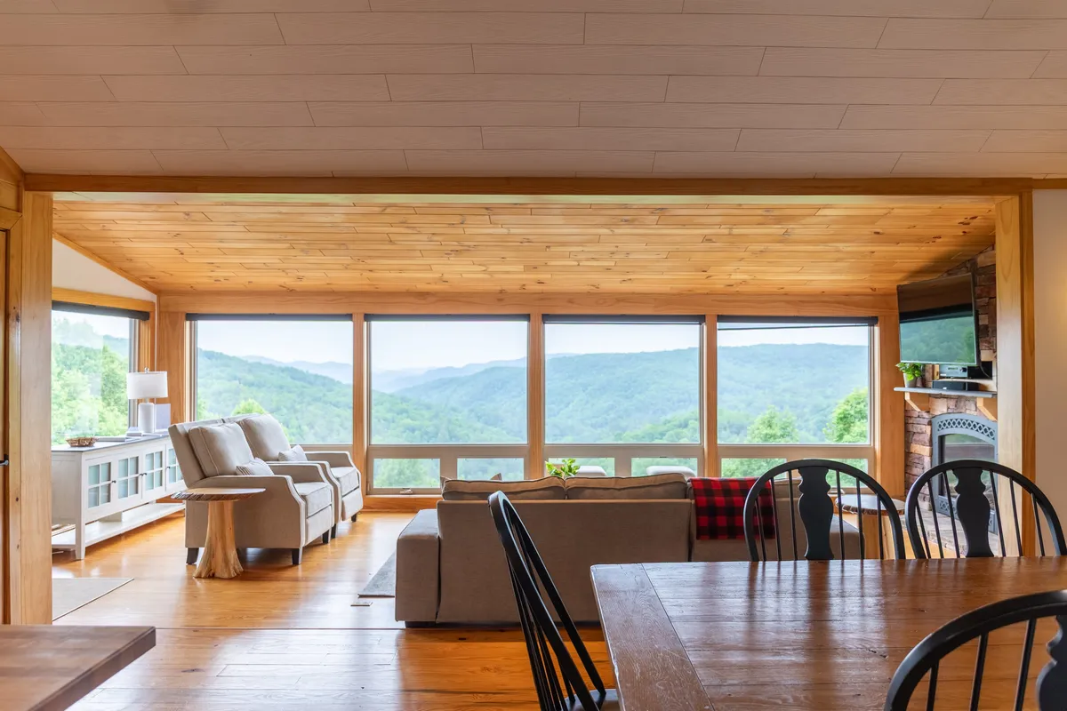 This image highlights an open-concept interior with large windows framing picturesque mountain views, a comfortable seating area, and a wooden dining table set for gatherings. The arrangement showcases the seamless connection between living, dining, and scenery.