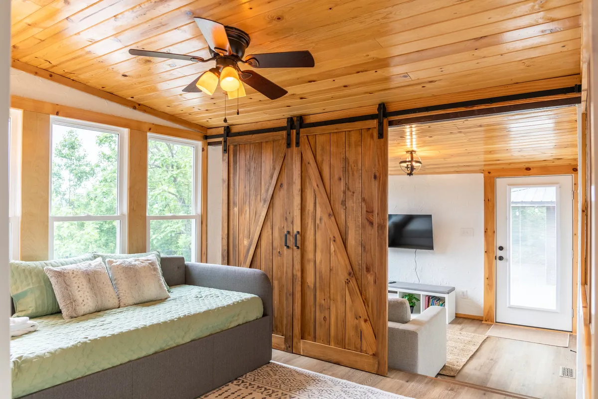 A playful and cozy nook with a daybed adorned with decorative pillows, flanked by barn doors, windows streaming in natural light, a ceiling fan overhead, and a TV on a stand. The setting feels intimate and versatile, ideal for lounging or sleeping.