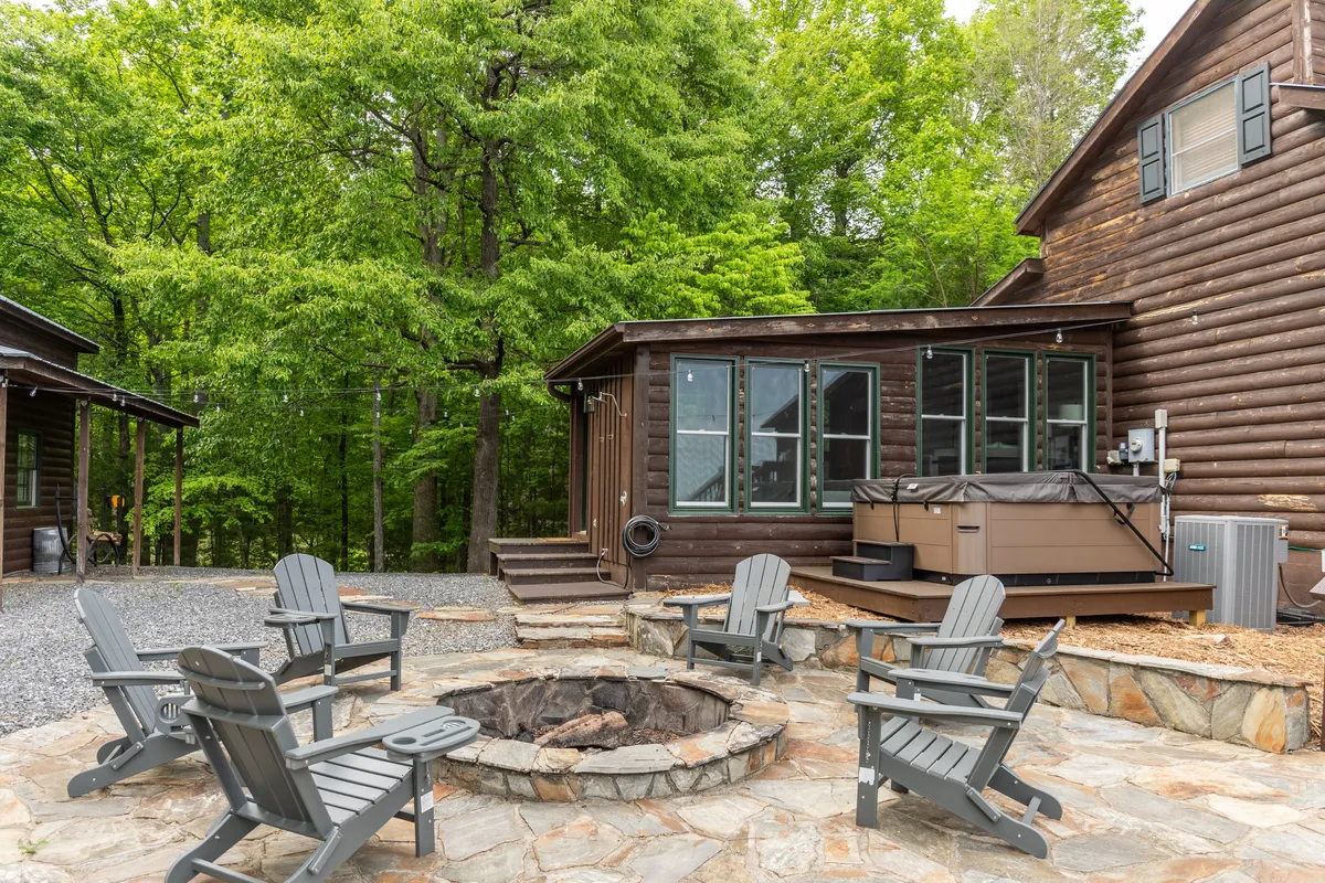An inviting outdoor scene featuring a fire pit surrounded by comfortable seating and a hot tub, set against a backdrop of trees and distant mountain views. The area is clearly designed for communal gatherings, relaxation, and socializing under the open sky.